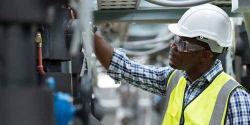 Technician wearing hard hat, goggles and a vest examining industrial equipment.
