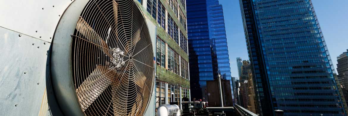 External HVAC fan on side of building in foreground with blue sky and office buildings in background.